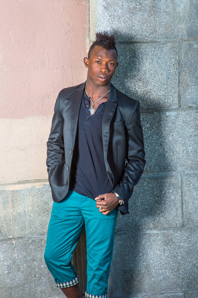 Dressing in a black blazer, a black shirt and green pants, a young black guy with mohawk hair is standing against the wall, confidently looking at you - Foto, Imagem
