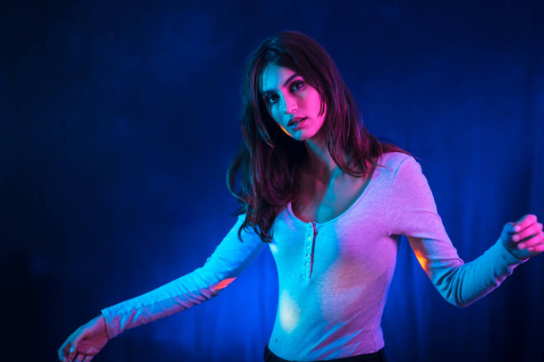 A young brunette Caucasian woman in a white t-shirt in a nightclub with a black background. Neon lights, dancing portrait - Fotó, kép
