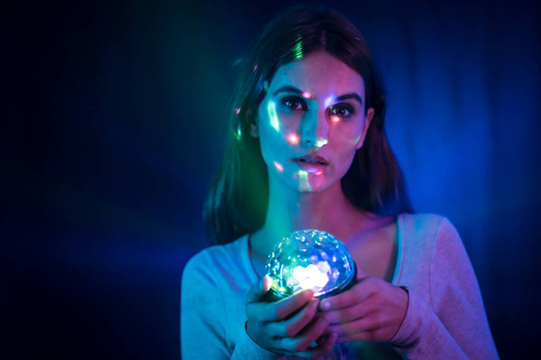 A young brunette Caucasian woman in a white t-shirt on a black background. Neon lights, illuminated with a disco light in hand - Foto, Imagen