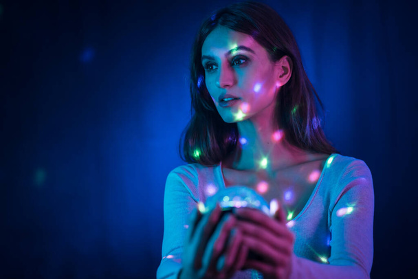 A young brunette Caucasian woman in a white t-shirt on a black background. Neon lights, illuminated with a disco light in hand, sweet look - Fotó, kép