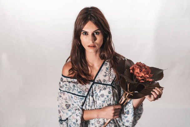 A young brunette Caucasian woman in a vintage floral dress on a white background. Portrait with a red flower in hand, with a sweet look in the studio - Foto, immagini