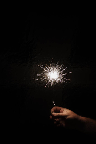 Рука с блеском на темном фоне. Празднование нового года с бенгальским огнем. Рука держит искру на темном фоне. Рождественское веселье Новогодняя концепция
. - Фото, изображение