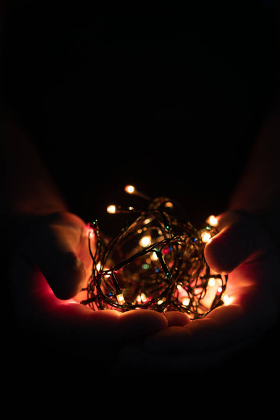 male hands holding a Christmas garland with lights on a dark background with copy space for text. Retro effect, close-up. Winter, postcard template - Foto, imagen