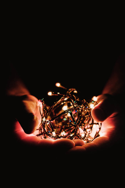 male hands holding a Christmas garland with lights on a dark background with copy space for text. Retro effect, close-up. Winter, postcard template - Foto, imagen