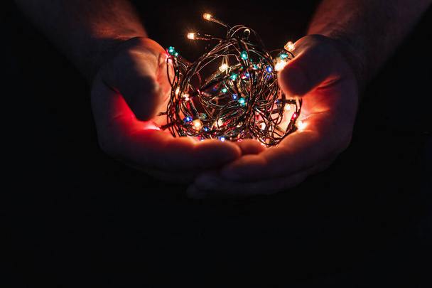 male hands holding a Christmas garland with lights on a dark background with copy space for text. Retro effect, close-up. Winter, postcard template - 写真・画像
