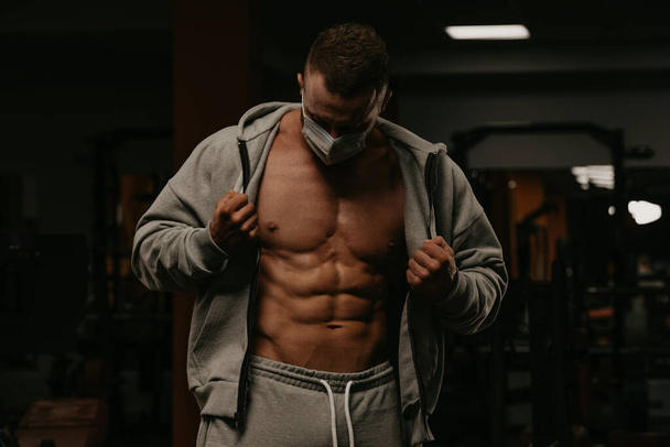 A bodybuilder in a face mask to avoid the spread of coronavirus is opening his zipped hoodie to demonstrate his athletic physique. A sporty guy in a surgical mask is posing after a workout in a gym. - Fotografie, Obrázek