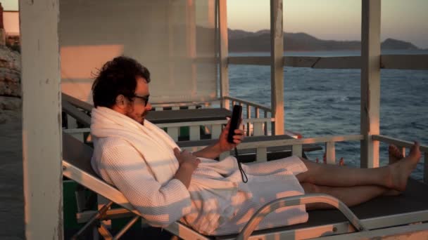 Fiatal szakállas férfi beszél videohívással az okostelefonján fekszik a napozóágyon a társalgóban. - Felvétel, videó