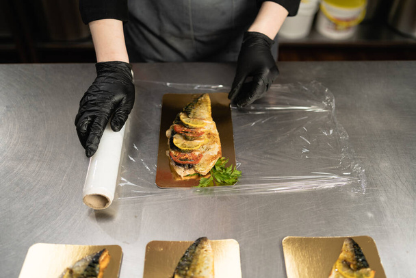 台所のレストランでテーブルの上に食べ物を保存するためにラップフィルムを使用して女性。食品包装用透明プラスチック製のリングフィルムのロール。焼きサバ。スペースのコピー. - 写真・画像
