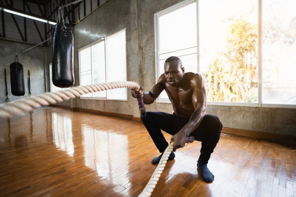 アフリカのスポーツマンはジムで戦闘ロープ機能訓練を行っている。力を行使しながらロープで波を作るトレーナーを決定しました。産業用ジムでバトルロープを使用して運動選手. - 写真・画像
