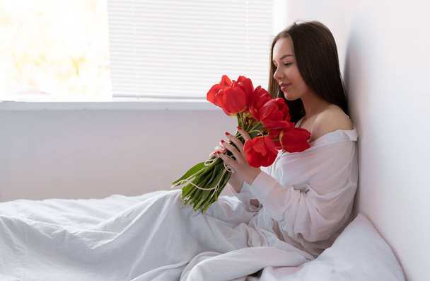 joven hermosa mujer por la mañana en el dormitorio se sienta con un gran ramo de tulipanes. flores a una mujer para el aniversario, el día de San Valentín, o simplemente así. Copiar espacio. - Foto, Imagen