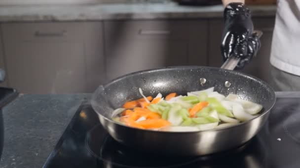 Chef en guantes negros cocinando verduras en sartén en la cocina del restaurante. Mezclar y remover los ingredientes en cámara lenta. Preparación de alimentos. Chef ocupado trabajando. Full hd - Metraje, vídeo