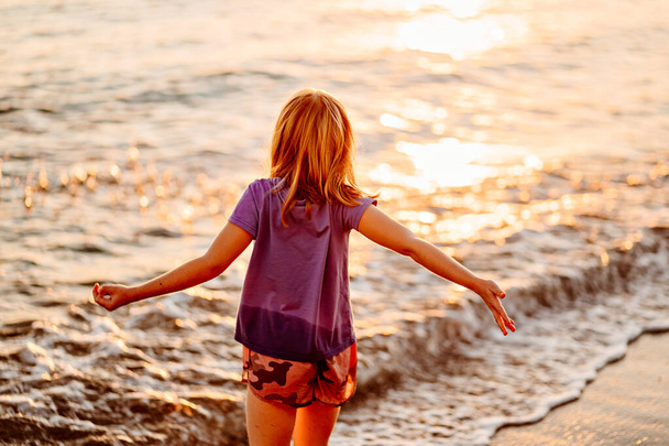 at sunset. kid girl walks along seashore, collects stones and throws into water - 写真・画像