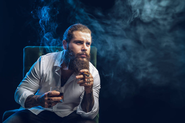 Brutal hipster bearded man smoke a cigar drinks whiskey, dressed in white shirt. Studio shot - 写真・画像