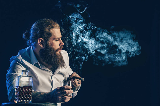 Handsome bearded guy smoke a cigar holds glass of whiskey, blows smoke. Studio shot - Zdjęcie, obraz