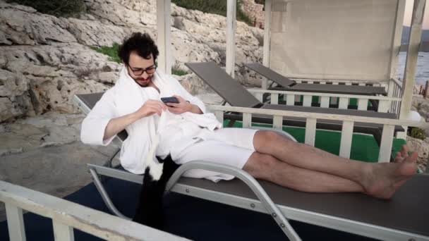 Joven caucásico barbudo hombre está descansando en sala de estar tumbado en la cama de sol y jugando con un gato - Imágenes, Vídeo