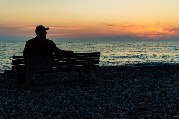 a man in a cap sits alone on a bench by the sea and looks to the right. relaxation by the sea during the evening sunset - Photo, Image