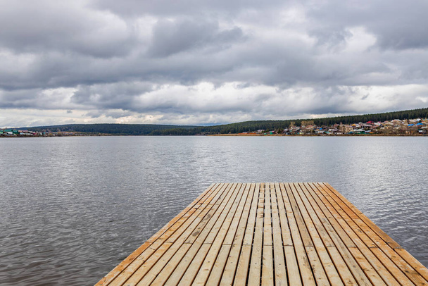 wooden pier on a pond under a cloudy tragic sky, in the distance a village and hills covered with forest - Photo, Image