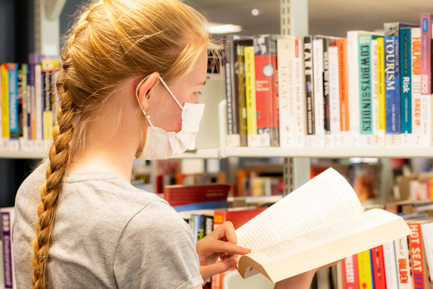 2021-12-02 Sydney, Austrlaia Adolescente con máscara facial con el libro en sus manos en la biblioteca durante la pandemia de COVID-19. Biblioteca escolar - Foto, Imagen