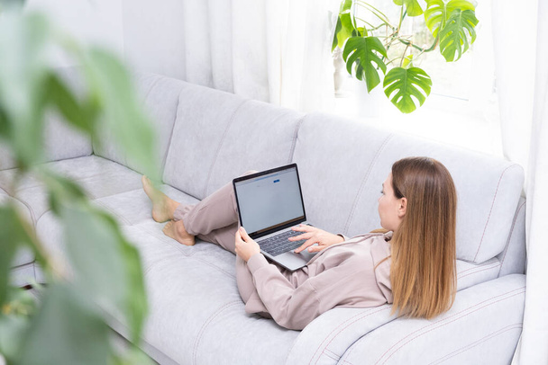 Νεαρή ή μεσήλικη γυναίκα κάθεται με φορητό υπολογιστή σε γκρι καναπέ στο γραφείο στο σπίτι με monstera φυτό. Έννοια του απομακρυσμένου χώρου εργασίας και εργασίας στο σπίτι. - Φωτογραφία, εικόνα