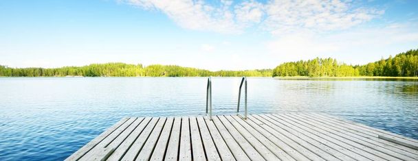 Wooden pier near the river close-up. Evergreen coniferous forest in the background. Clear blue sky, reflections on the water. Idyllic summer landscape. Eco tourism, recreation theme. Finland - Photo, Image