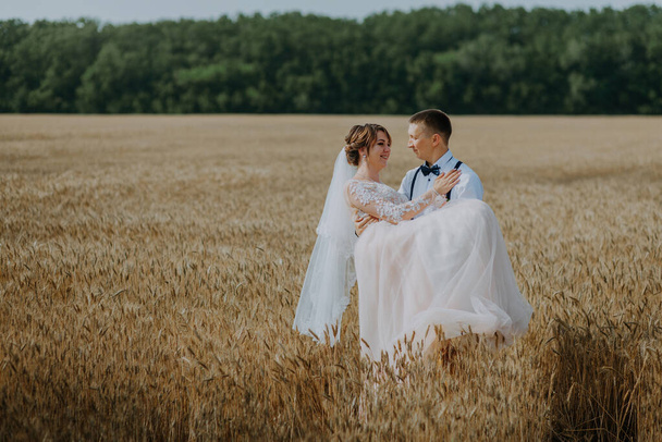 fashionable and happy wedding couple at wheat field at sunny day. Bride and groom kissing in a wheat field. Young beautiful wedding couple hugging in a field with grass eared. - Zdjęcie, obraz