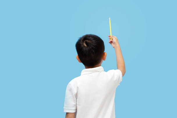 Niño hoding lápiz y apuntando hacia arriba con camisa blanca, vista trasera, sobre fondo azul - Foto, Imagen