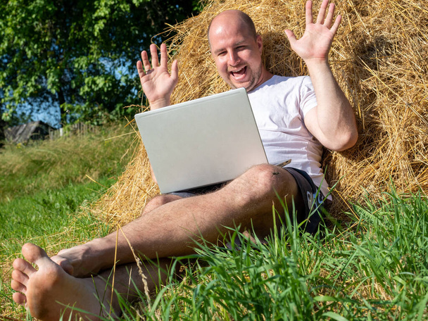 a middle-aged man lying on a yellow haystack with a gray laptop in his hands. Remote work, training. Rural area. Joyful facial expression, waving his arms, emotions - 写真・画像