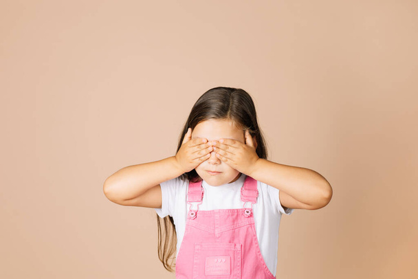 Close up portrait rebellious kid closing eyes with hands not see others with closed mouth not talk with others wearing bright pink jumpsuit and white t-shirt on beige background - Fotó, kép