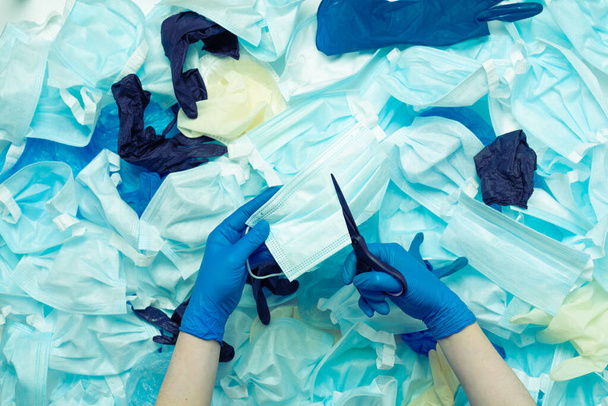 Hands with scissors cut through the medical mask in two against the background of medical waste. - Photo, image