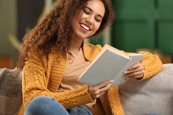 Молода афро-американська жінка в жовтому кардиганському читанні на дивані вдома. - Фото, зображення