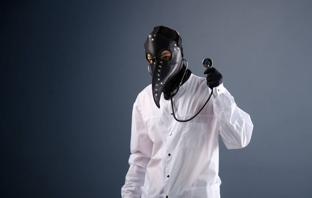 a doctor in a dressing gown wearing a plague doctor mask with a stethoscope in his hands - Фото, изображение