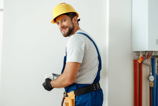 Selbstbewusster Elektriker mit Mütze und Schutzhandschuhen lächelt in die Kamera, während er Steckdose in einem Raum installiert - Foto, Bild