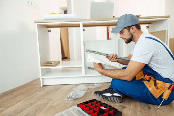 Rušný údržbář v montérkách sedí na podlaze a čtení manuál při montáži kuchyňské skříňky v bytě - Fotografie, Obrázek