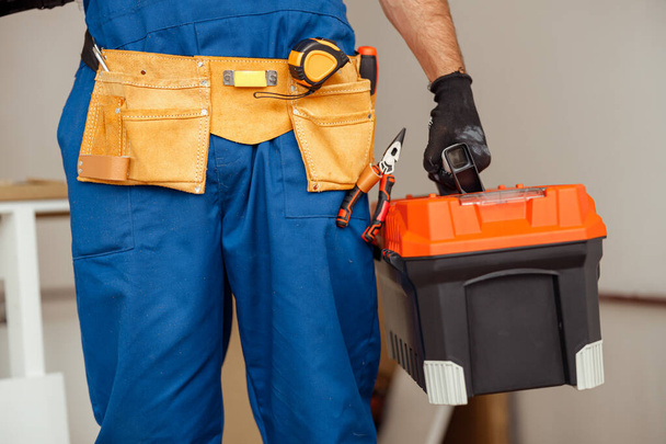 Closeup shot of contractor worker in overalls holding toolbox while standing indoors - Photo, image