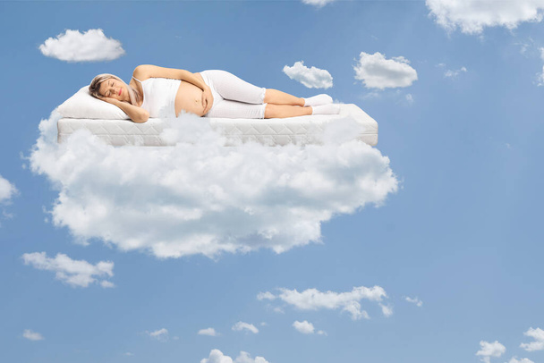 Pregnant woman in pajamas sleeping on a mattress and floating on clouds in the sky - Fotoğraf, Görsel