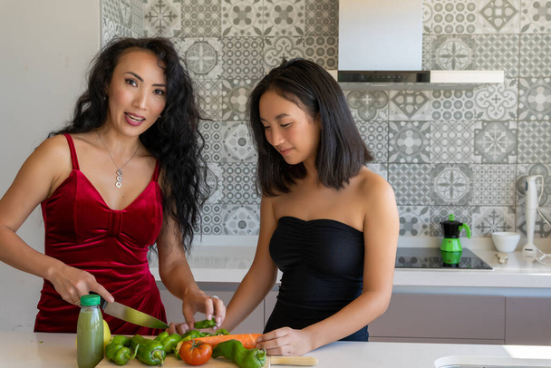dos mujeres cocinando en la cocina vistiendo vestidos, uno de ellos cortando verduras, fondo con dibujos horizontales - Foto, imagen