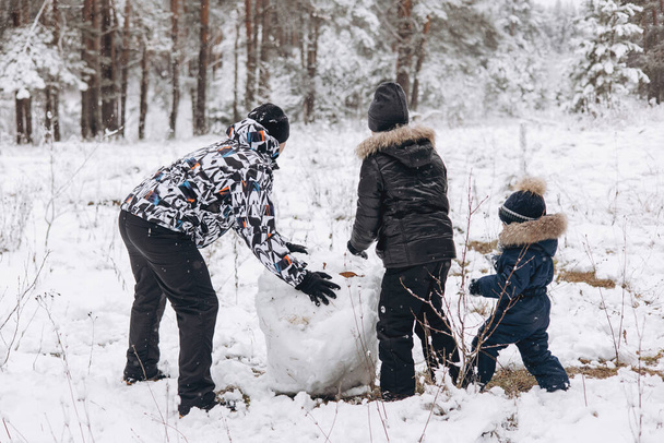 Happy father and sons rolling big snowball together for snowman in snowy winter forest. Man Joyful teenager and little boy playing and having fun on family walking. Wintertime activity outdoors. - Photo, Image