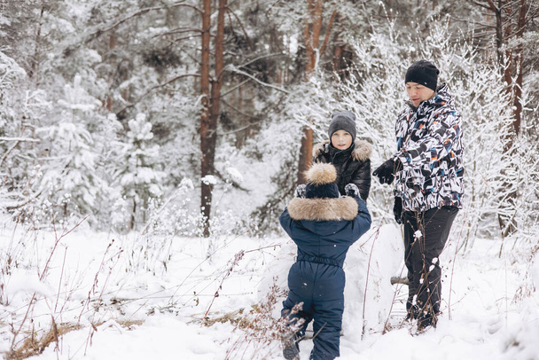 Happy father and sons rolling big snowball together for snowman in snowy winter forest. Man Joyful teenager and little boy playing and having fun on family walking. Wintertime activity outdoors. - Foto, Imagem