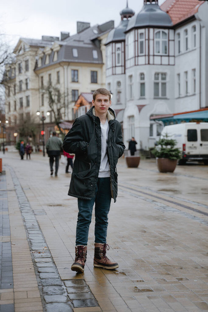 ένας νεαρός άνδρας στέκεται στη μέση του δρόμου, κρατά τα χέρια του στις τσέπες του, κοιτάζει την κάμερα με ένα σοβαρό πρόσωπο, σε ένα μαύρο μπουφάν χειμώνα, κρύα υγρή πόλη - Φωτογραφία, εικόνα