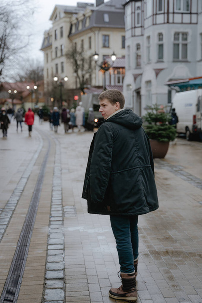 un adolescente camina por las calles vacías de la ciudad, dando la vuelta y mirando hacia un lado, con una chaqueta de invierno negro, en los murciélagos altos de invierno - Foto, imagen
