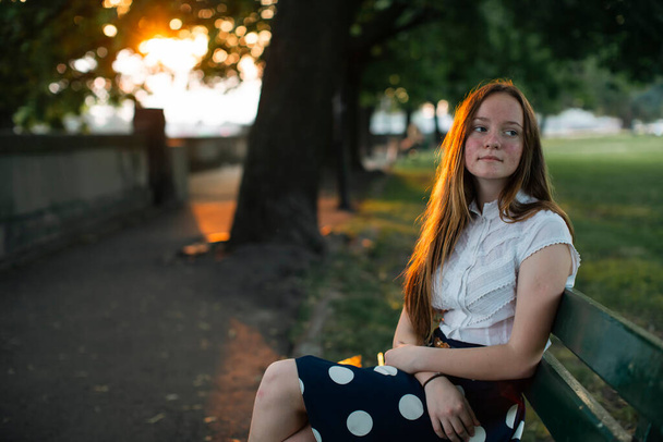 Ένα κορίτσι με χρυσά μαλλιά κάθεται σε ένα καλοκαιρινό δρομάκι της πόλης πριν το ηλιοβασίλεμα. - Φωτογραφία, εικόνα
