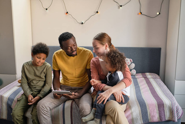 Счастливая молодая семья из четырех человек сидит на двуспальной кровати, смотрит онлайн-видео и обсуждает его - Фото, изображение