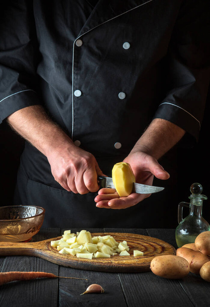 Professional chef cuts raw potatoes into pieces with a knife before preparing breakfast or dinner. Close-up of a cook hands while working in a restaurant kitchen. - Foto, Imagem