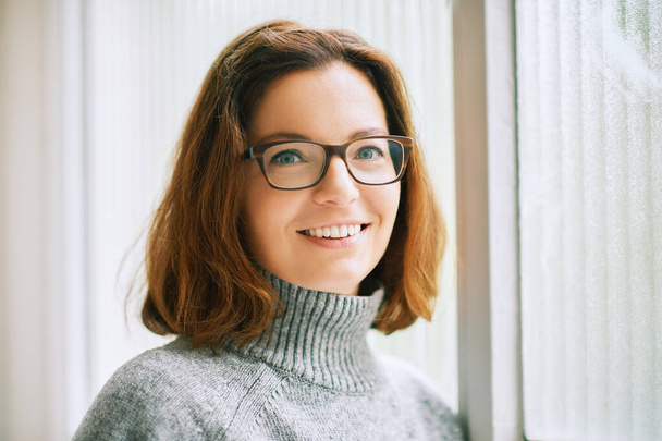 Interior portrait of beautiful 40 year old woman relaxing next to window, wearing warm grey pullover and glasses - Photo, Image