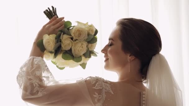 Невеста в белом платье, стоящая у окна с цветочным букетом, свадебные утренние приготовления дома - Кадры, видео