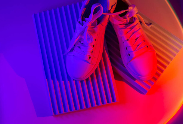 Divatos fehér cipők geometria pódiumon elvont fényes háttérrel. Neonfények alkalmi cipőn. Violet és piros gradiens lámpa. Minimalizmus, a 90-es, 80-as évek fogalma. - Fotó, kép