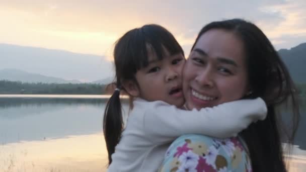 Cute little girl and her mother hugging in love playing on a summer day by the lake at sunset. Happy family spending time together on vacation. - Filmagem, Vídeo