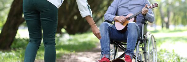 Disabled man in wheelchair plays guitar in park passers-by give money. - Zdjęcie, obraz