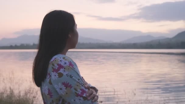 Genç Asyalı kadın gün batımında göl kenarında yalnız hissederek kollarını kavuşturuyor. Yanlız bir kadın kışın göl kenarında temiz hava soluyor.. - Video, Çekim