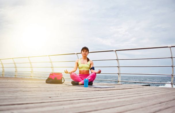 Młoda skoncentrowana kobieta siedząca na macie do jogi w lotosowej pozie na plaży. Wewnętrzna harmonia - Zdjęcie, obraz